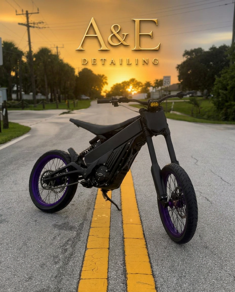 The A&E Detailing logo in gold lettering above a matte black e-bike with purple rims, parked on a quiet road at sunset